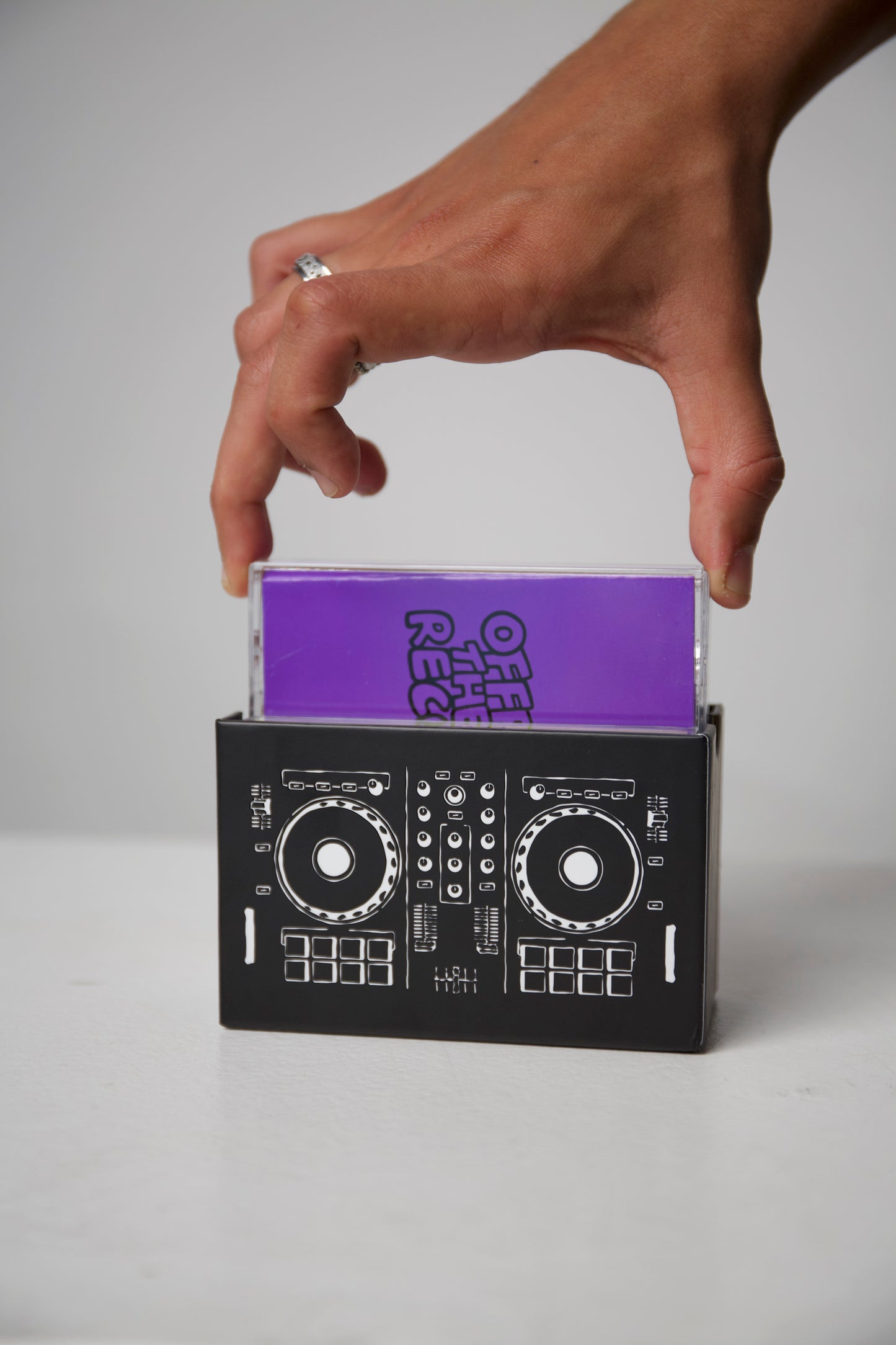 Hand holding a purple book titled 'Off the Record' inside a black DJ console-shaped box on a gray background.