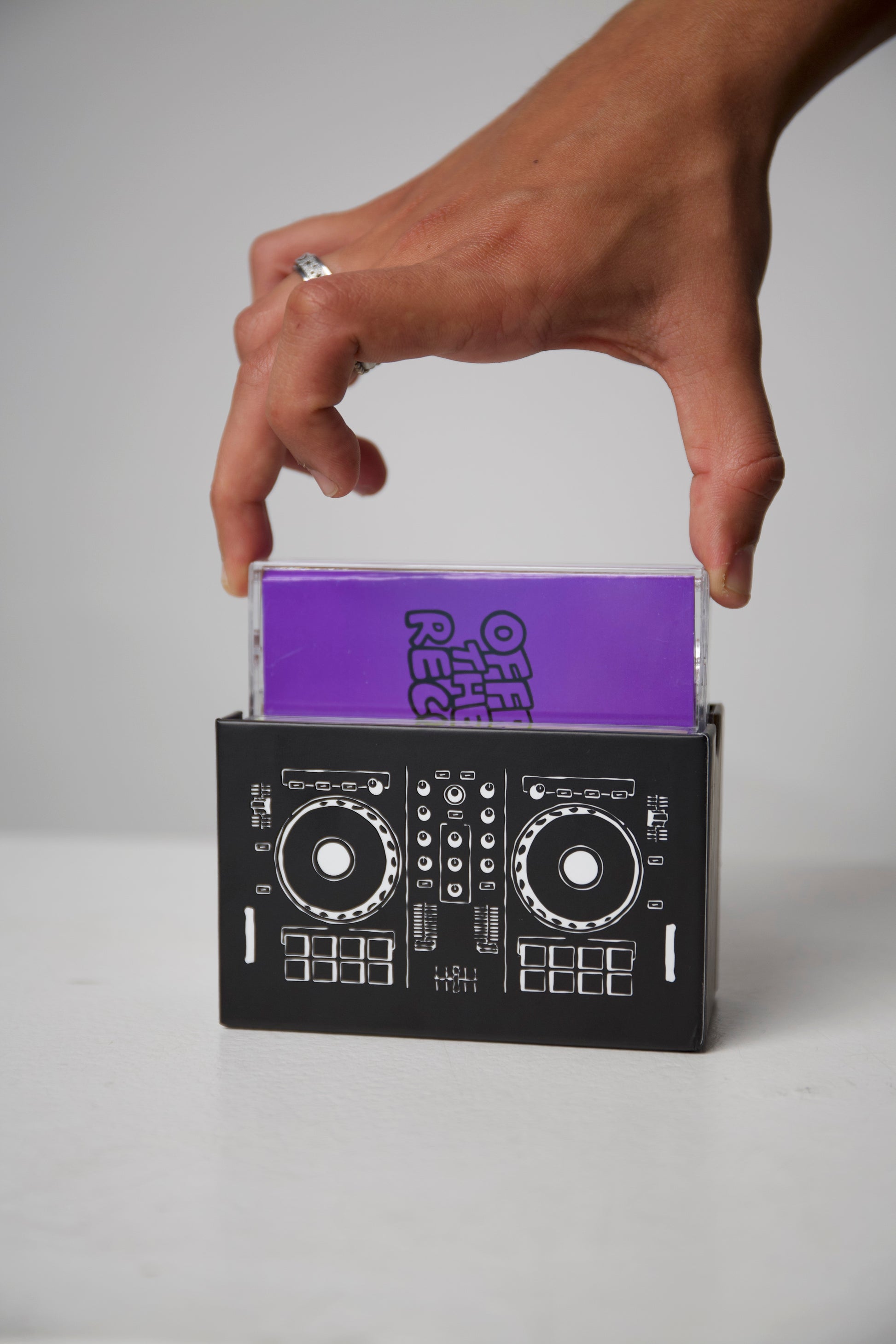 Hand holding a purple book titled 'Off the Record' inside a black DJ console-shaped box on a gray background.